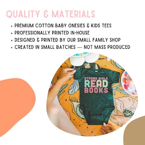 a person's hands holding a baby onesie with the text "STRONG GIRLS READ BOOKS" printed on it, placed on a floral patterned blanket.