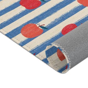 May include: A rectangular area rug featuring a pattern of red apples with green leaves set against a background of thick, textured blue stripes on a cream-colored fabric. The rug is partially rolled, revealing a gray, woven backing material.