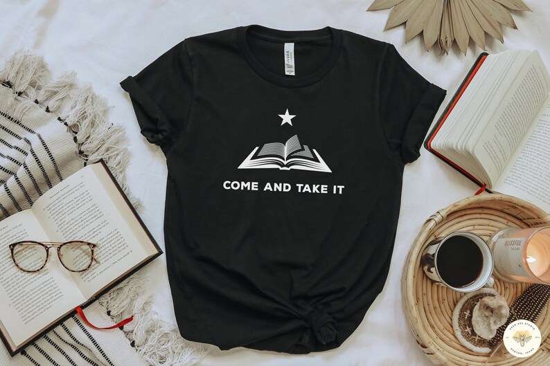 May include: A black short-sleeved t-shirt featuring a white graphic of an open book with a single star positioned above it. Below the book, the text reads "COME AND TAKE IT" in white capital letters. The shirt has a crew neckline and short sleeves.