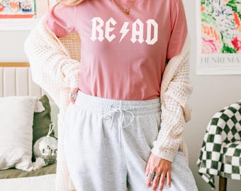 May include: A dusty rose short-sleeved t-shirt featuring the white text READ with a lightning bolt replacing the letter A. The shirt is worn with a cream-colored open-front knit cardigan and light gray drawstring sweatpants.