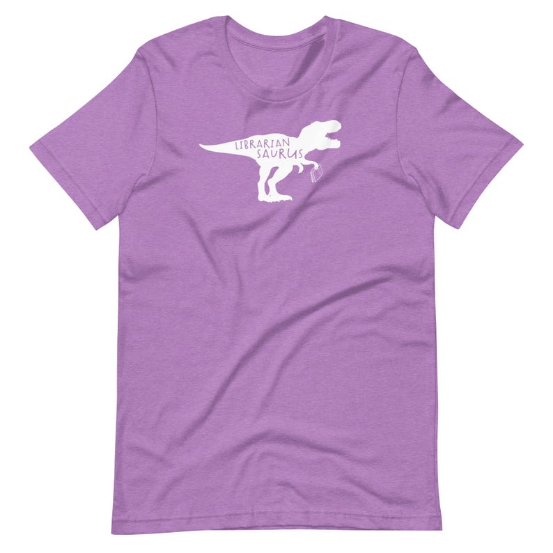 May include: A lilac-colored t-shirt featuring a white graphic of a dinosaur silhouette holding a coffee cup. The text "Librarian Saurus" is printed above the dinosaur. The shirt has a crew neck and short sleeves.