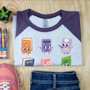 May include: A light gray raglan t-shirt with dark purple sleeves featuring a graphic print of six anthropomorphic books labeled History, Fantasy, Cooking, Poetry, Romance, and Mystery. The shirt has a crew neckline and a visible interior brand tag.