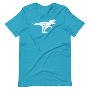 May include: A turquoise t-shirt with a white graphic of a Tyrannosaurus Rex holding a book. The text "Librarian Sauras" is above the dinosaur. The shirt is made of a soft material.