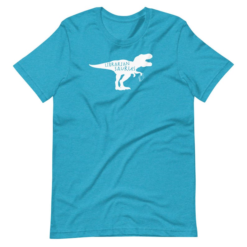 May include: A turquoise t-shirt with a white graphic of a Tyrannosaurus Rex holding a book. The text "Librarian Sauras" is above the dinosaur. The shirt is made of a soft material.