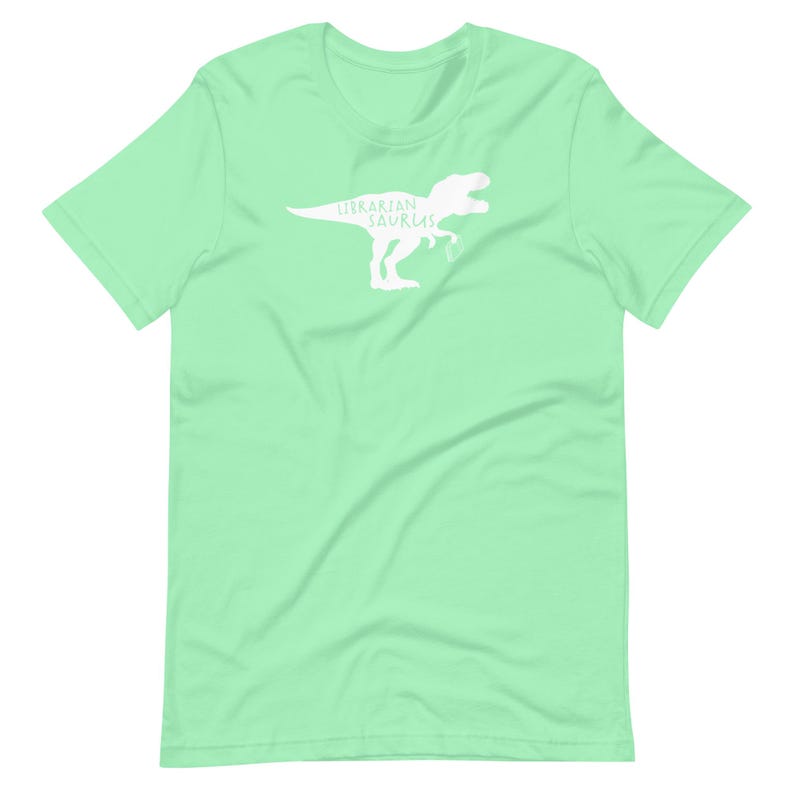 May include: A light green t-shirt featuring a white silhouette of a Tyrannosaurus Rex with the words "Librarian Saurus" printed on it. The shirt is made of a soft material and has a crew neck.