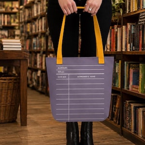 May include: A purple tote bag with two mustard yellow straps, featuring a white graphic print of a library checkout card. The print includes the text AUTHOR, TITLE, DATE DUE, and BORROWER'S NAME above a series of horizontal lines for entries.