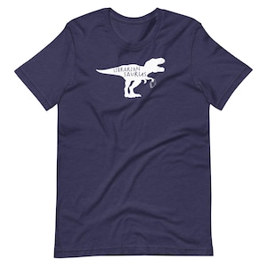 May include: Navy blue t-shirt featuring a white graphic of a dinosaur holding a book, with the text "Librarian Saurus". The shirt is made of a soft material and has short sleeves and a crew neck.