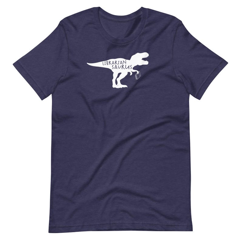 May include: Navy blue t-shirt featuring a white graphic of a dinosaur holding a book, with the text "Librarian Saurus". The shirt is made of a soft material and has short sleeves and a crew neck.