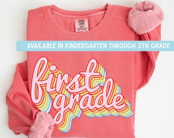 Może przedstawiać: Koralowa bluza z okrągłym dekoltem z napisem "first grade" wykonanym wielokolorową czcionką w stylu retro. Bluza posiada metkę Comfort Colors przy kołnierzu oraz napis "AVAILABLE IN KINDERGARTEN THROUGH 5TH GRADE" umieszczony na środku.
