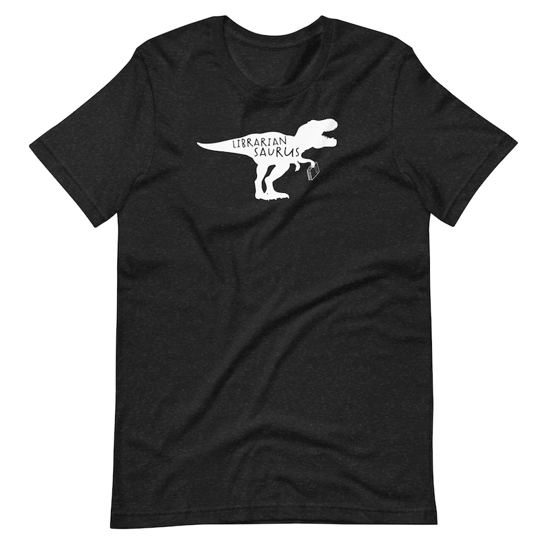 May include: Dark gray t-shirt featuring a white graphic of a Tyrannosaurus Rex holding a coffee mug. The text "Librarian Saurus" is above the dinosaur. The shirt is made of a soft material.