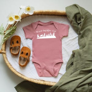 May include: A pink baby bodysuit with the text "Second Breakfast Club" in white, and a silhouette design. The bodysuit is next to brown suede baby shoes and a green muslin blanket, in a woven basket.