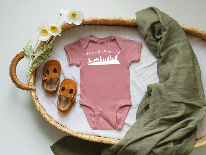 May include: A pink baby bodysuit with the text "Second Breakfast Club" in white, and a silhouette design. The bodysuit is next to brown suede baby shoes and a green muslin blanket, in a woven basket.