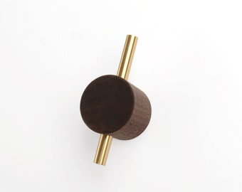 May include: A modern wall hook featuring a dark brown, cylindrical wooden body with a brass rod extending through it. The hook is designed for hanging items and is set against a plain white background.