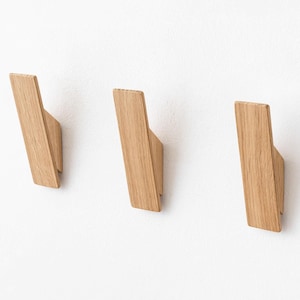 May include: Three wooden wall hooks in a modern design. Each hook is made of light-coloured wood and has a slanted, angular shape. The hooks are mounted on a white wall, providing a minimalist aesthetic.