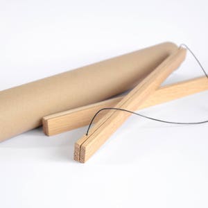 Puede incluir: Un kit para colgar pósteres con un tubo de cartón marrón y barras de madera. Las barras de madera son de color marrón claro y están conectadas con una cuerda negra. El tubo tiene un interior blanco.