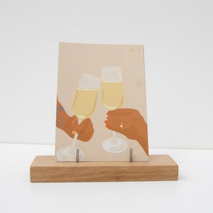Puede incluir: Ilustraci&oacute;n de tarjeta de felicitaci&oacute;n que representa dos copas de champ&aacute;n brindando, sostenidas por manos. La tarjeta se muestra en un soporte de madera. La ilustraci&oacute;n tiene un fondo beige con las letras "A&T" en la esquina superior derecha.