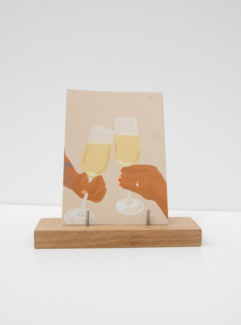Puede incluir: Ilustraci&oacute;n de tarjeta de felicitaci&oacute;n que representa dos copas de champ&aacute;n brindando, sostenidas por manos. La tarjeta se muestra en un soporte de madera. La ilustraci&oacute;n tiene un fondo beige con las letras "A&T" en la esquina superior derecha.