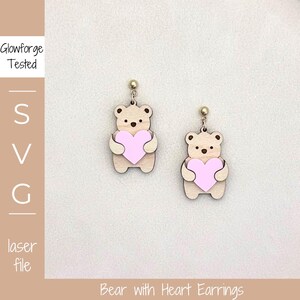 Pode incluir: Um par de brincos de urso de madeira, cada um segurando um coração rosa. Os brincos têm pinos dourados e são cortados a laser. O texto "Glowforge Tested" e "SVG laser file" é visível.
