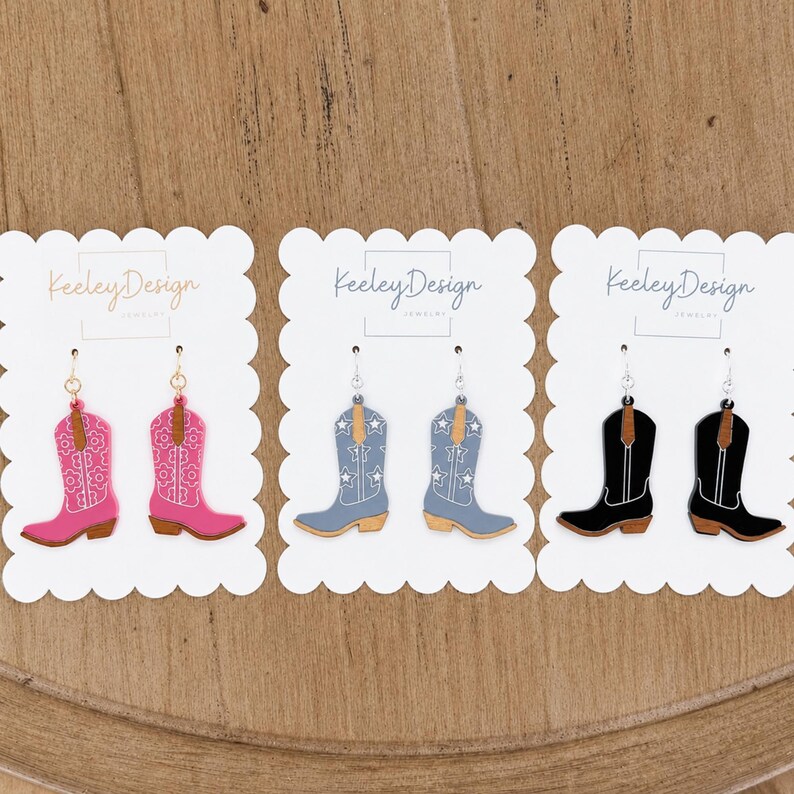 May include: Three pairs of dangling earrings shaped like cowboy boots, displayed on white scalloped cards labeled Keeley Design. The first pair is pink with white floral patterns, the second is blue with white stars, and the third is black with white outlines.