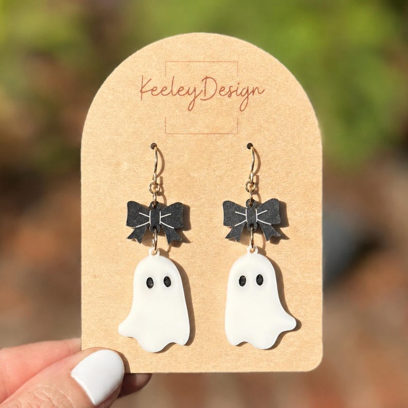May include: A pair of dangle earrings on a tan display card labeled Keeley Design. Each earring features a black bow with white line details attached to a white ghost charm with two black oval eyes. The earrings hang from gold-toned metal hooks.
