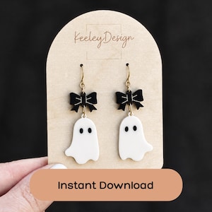 May include: A pair of dangle earrings featuring white ghost charms with black eyes and mouths, topped with black bows. The earrings hang from gold-toned metal hooks and are displayed on a light-colored arched card labeled Keeley Design.