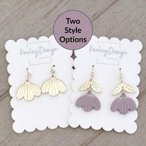 May include: Two pairs of earrings on white scalloped cards. One pair is gold-colored, flower-shaped, and the other is purple with gold leaf accents. The text "Two Style Options" is in a circle above the earrings. The brand name "Keeley Design Jewelry" is printed on the cards.