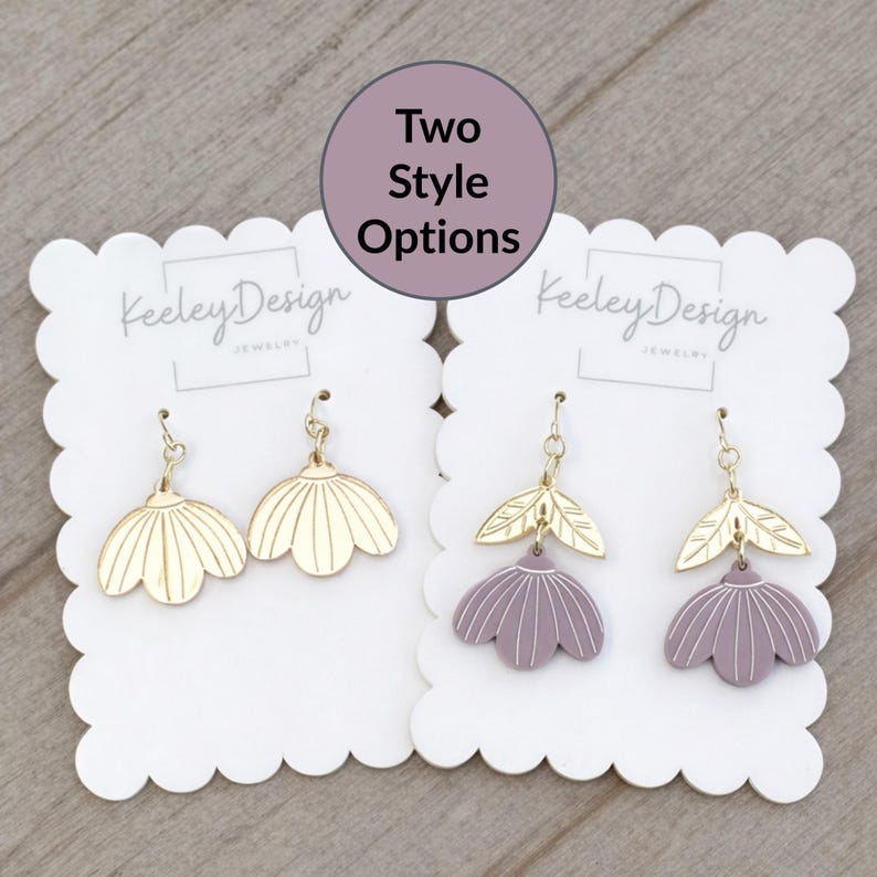 May include: Two pairs of earrings on white scalloped cards. One pair is gold-colored, flower-shaped, and the other is purple with gold leaf accents. The text "Two Style Options" is in a circle above the earrings. The brand name "Keeley Design Jewelry" is printed on the cards.