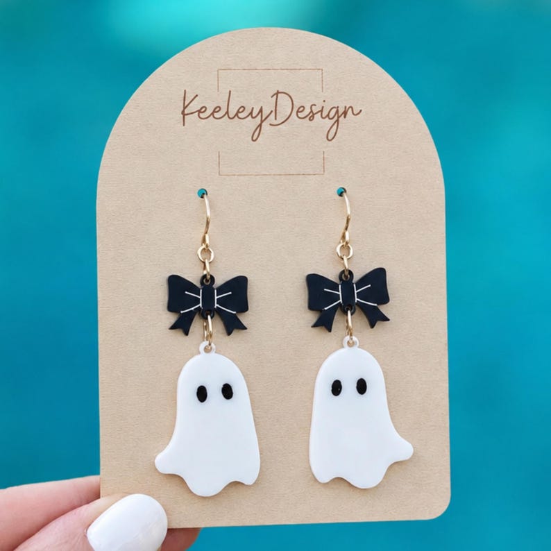 May include: A pair of dangle earrings on a tan display card labeled Keeley Design. Each earring features a black bow with white line details attached to a white ghost charm with two black oval eyes, suspended from gold-toned metal earring hooks.