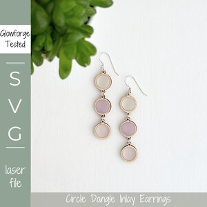 May include: A pair of dangle earrings with three circles each. The circles are made of wood and have a white and light purple inlay. The earrings are hanging from silver hooks.