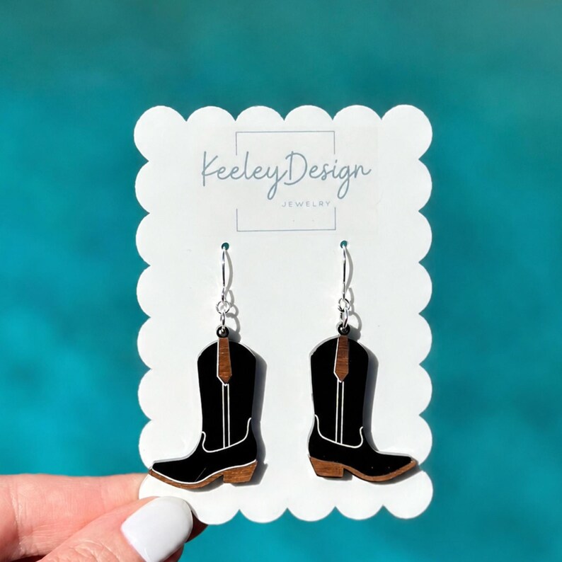 May include: A pair of dangle earrings shaped like cowboy boots on a white scalloped display card. The boots are black with brown wood-tone accents at the top and bottom, featuring white line details. The card displays the text Keeley Design Jewelry.