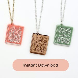 May include: Three necklaces featuring book-shaped pendants on silver-toned chains. The left pendant is coral with white floral line art, the centre is natural wood with dark floral line art, and the right is sage green with white floral line art and the text Wildflowers.