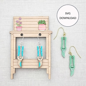 May include: Wooden earrings in the shape of garden shears, with turquoise handles and silver accents, hanging from gold-colored ear wires. A matching pair of earrings is displayed next to a small wooden potting bench with potted plants. The image includes the text "SVG DOWNLOAD".