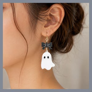 May include: A dangle earring featuring a white ghost charm with two black oval eyes, suspended from a small black bow with white line details. The charm is attached to a gold-toned metal hook that secures the piece to the earlobe.