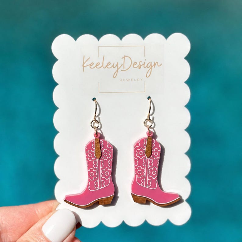 May include: A pair of pink cowboy boot earrings featuring a white floral pattern and brown accents. The earrings are attached to a white scalloped display card with the text Keeley Design JEWELRY printed in the center in a brown script font.