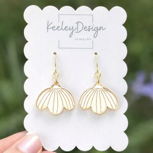 May include: A pair of gold-colored earrings with a cream-colored, flower-shaped design. The earrings are displayed on a white card with the text "Keeley Design JEWELRY". The background is a soft, blurred green.
