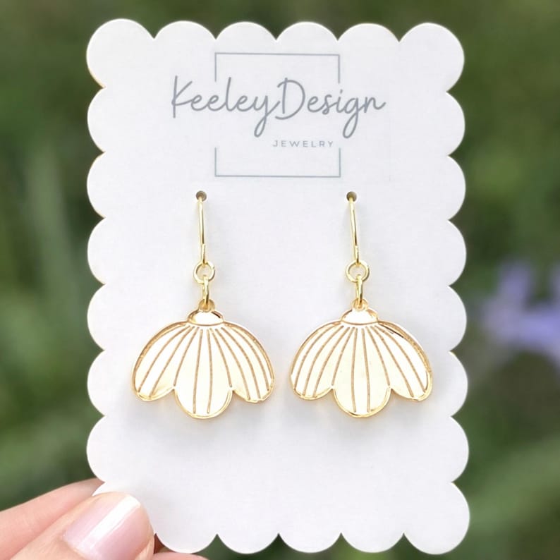 May include: A pair of gold-colored earrings with a cream-colored, flower-shaped design. The earrings are displayed on a white card with the text "Keeley Design JEWELRY". The background is a soft, blurred green.
