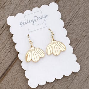 May include: Gold-colored earrings in a floral design, hanging from a gold-colored hook. The earrings are displayed on a white scalloped card with the text "Keeley Design JEWELRY". The background is a light brown wooden surface.