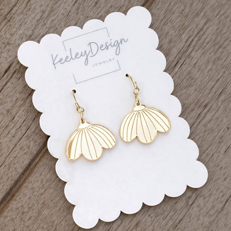May include: Gold-colored earrings in a floral design, hanging from a gold-colored hook. The earrings are displayed on a white scalloped card with the text "Keeley Design JEWELRY". The background is a light brown wooden surface.