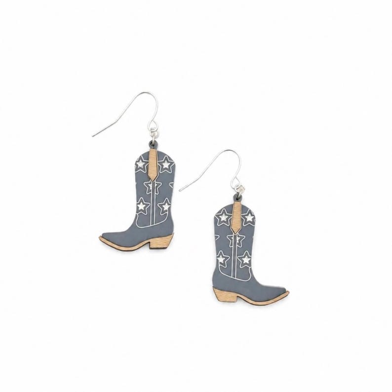 May include: A pair of dangle earrings shaped like cowboy boots. The boots are matte gray with white star outlines and white stitching details. The pull straps and heels are a natural light tan wood color. Each earring hangs from a silver hook.