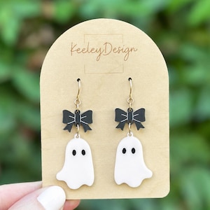 May include: A pair of dangle earrings featuring white ghost charms with black eyes and mouths, suspended from black bows with white line details. The earrings are attached to a tan arched display card with the text Keeley Design printed on it.