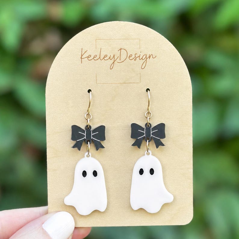 May include: A pair of dangle earrings featuring white ghost charms with black eyes and mouths, suspended from black bows with white line details. The earrings are attached to a tan arched display card with the text Keeley Design printed on it.