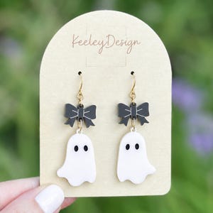 May include: A pair of dangle earrings featuring a white ghost charm with black eyes suspended from a black bow charm with white line details. The earrings are attached to a beige arched display card with the text KeeleyDesign printed in brown.