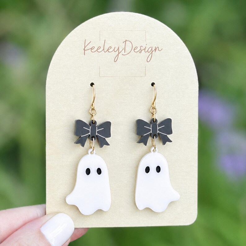 May include: A pair of dangle earrings featuring a white ghost charm with black eyes suspended from a black bow charm with white line details. The earrings are attached to a beige arched display card with the text KeeleyDesign printed in brown.