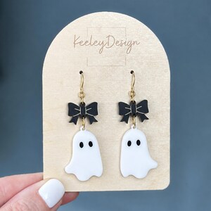 May include: A pair of dangle earrings featuring white ghost charms with black eyes and black bows attached to gold-toned metal hooks. The earrings are displayed on a light-colored wooden card with the text Keeley Design printed in black.
