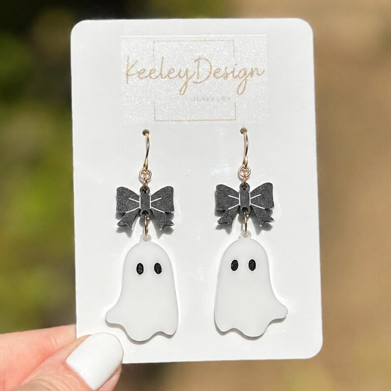 May include: A pair of dangle earrings on a white display card labeled Keeley Design. Each earring features a black bow charm attached to a gold-toned hook, with a white ghost pendant hanging below. The ghosts have two black oval eyes.