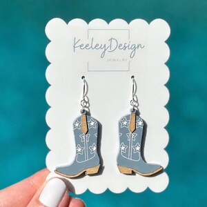 May include: A pair of dangle earrings shaped like cowboy boots in a light blue color with white star patterns and natural wood-toned accents. The earrings are displayed on a white scalloped card featuring the text Keeley Design Jewelry.