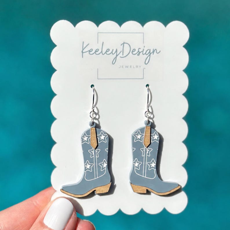 May include: A pair of dangle earrings shaped like cowboy boots in a light blue color with white star patterns and natural wood-toned accents. The earrings are displayed on a white scalloped card featuring the text Keeley Design Jewelry.