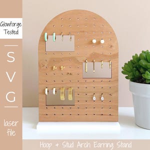 Puede incluir: Un soporte para pendientes de madera con forma de arco y base blanca. El soporte tiene un diseño de tablero perforado con agujeros para sujetar los pendientes. El soporte muestra una variedad de pendientes, incluyendo aros, tachuelas y pendientes colgantes.