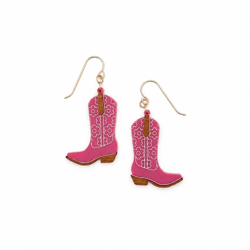 May include: A pair of drop earrings shaped like cowboy boots. The boots are pink with a white floral pattern and brown wood-tone accents at the top and heel. Each earring features a gold-toned metal hook for attachment to the earlobe.