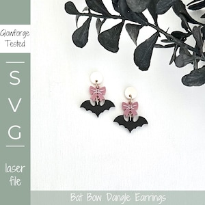 May include: Black bat-shaped dangle earrings with pink bows and white circular tops. The earrings are laser cut and feature the text "Bat Bow Dangle Earrings". The image also includes the text "Glowforge Tested" and "SVG laser file".